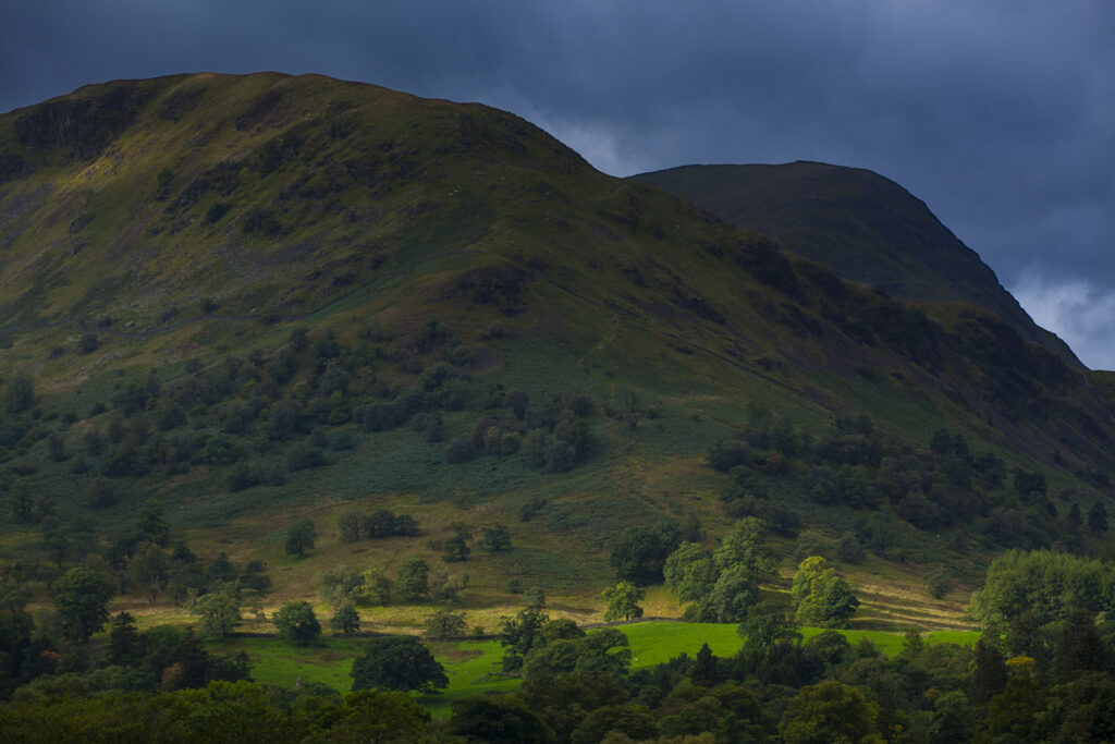 England, Cumbria, Lake District National Park. A soft shaft of light pieces a storm filled sky to highlight trees and fields on the lower fells near Glenridding in Patterdale.