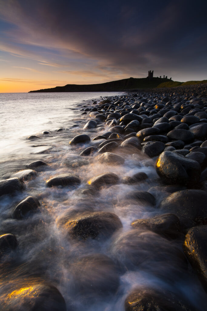 England, Northumberland, Embleton Bay. Waves crash againt the sea scultured rocks dominating the coastline of Embleton Bay, overlooked by the dramatic ruins of Dunstanburgh Castle.