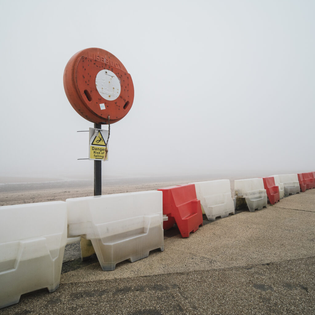 England, Northumberland, Blyth. Red & white safety barriers and lifebuoy on South Blyth Sea Front, photographed on a foggy morning.